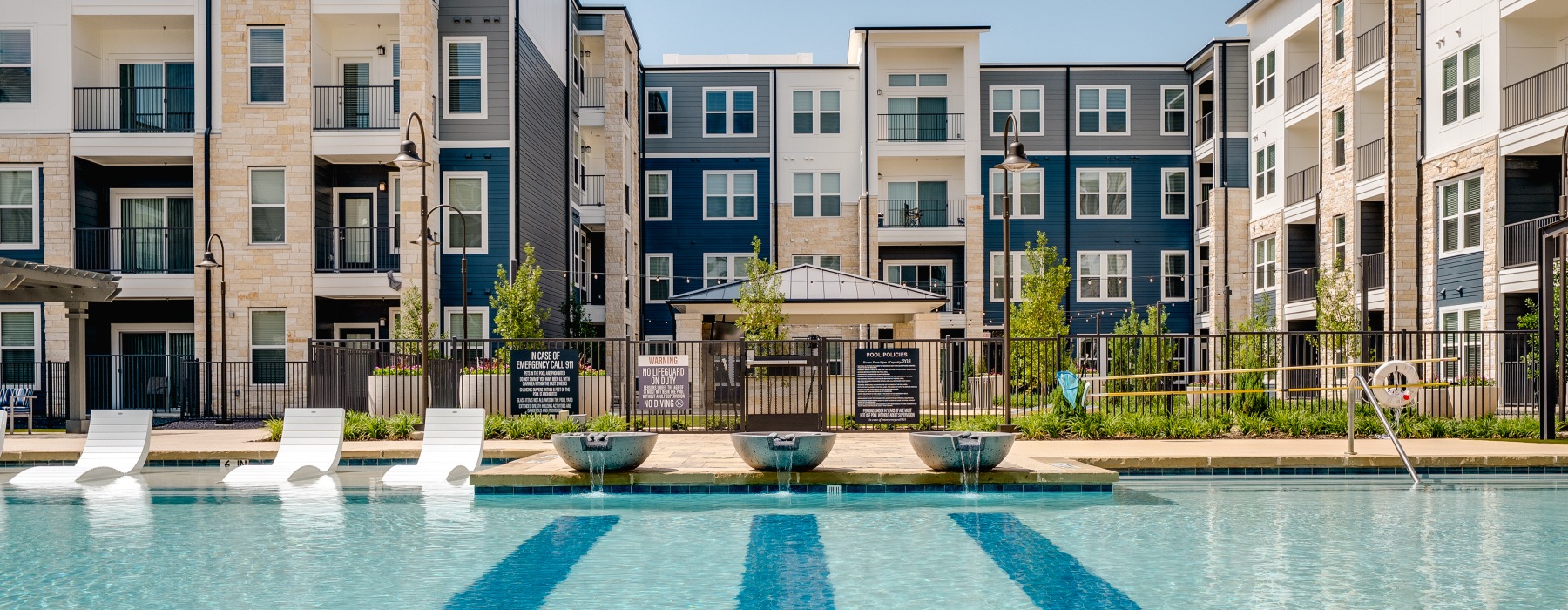 Sparkling pool with in water lounge chairs, green landascaing and the building in the  background